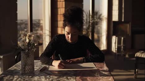 Young-sophisticated-female-artist-drawing-with-a-pen,-siting-by-the-table-in-art-studio-with-wide-panoramic-windows-on-the-background.-Inspiration,-full-concentration.