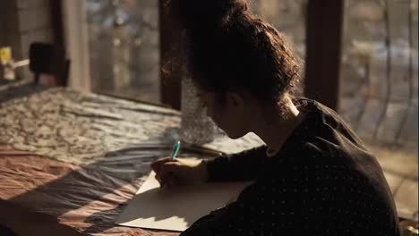 Lovely-female-artist-with-her-curly-hair-pulled-in-a-bun-sitting-by-the-table-near-panoramic-window-in-an-art-studio-with-minimalistic-interior-and-drawing-with-a-pen-on-paper