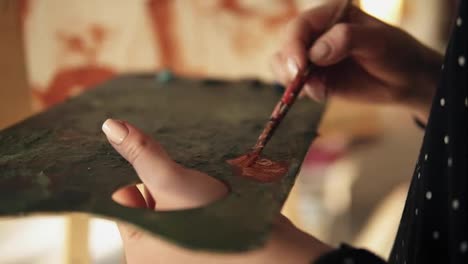 Close-up-slow-motion-footage-of-accurately-manicured-artist-hand-spreading-oil-paint-on-the-palette-and-adding-more-color-to-the-painting-on-easel