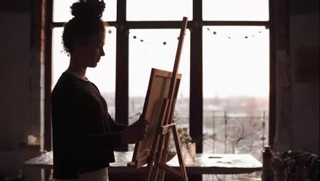 Young-inspired-professional-female-artist-puts-cancas-on-easel,-takes-palette-with-paint-on-it-and-starts-drawing.