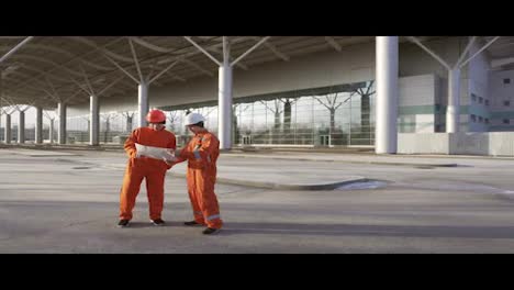 Bauarbeiter-In-Orangefarbenen-Uniformen-Und-Helmen-Schauen-Sich-Gemeinsam-Die-Pläne-An.-Gebäude-Im-Hintergrund
