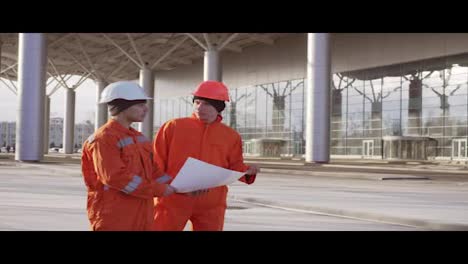 Two-construction-workers-in-orange-uniform-and-hardhats-examining-the-constructed-building-together.-Teamwork-concept