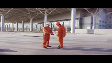 Two-construction-workers-in-orange-uniform-and-hardhats-meeting-each-other-at-the-bulding-object-and-examining-the-constructed-building-together.-Teamwork-concept