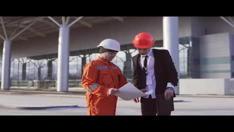 Young-architect-in-a-black-suit-with-tie-examining-the-building-object-with-construction-worker-in-orange-uniform-and-helmet.-They-meeting-each-other-at-the-bulding-object-and-shaking-hands.