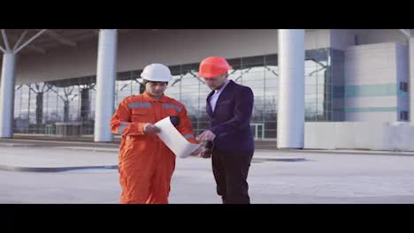 Young-architect-in-a-black-suit-examining-the-building-object-with-construction-worker-in-orange-uniform-and-helmet.-They-meeting-each-other-at-the-bulding-object-and-shaking-hands.