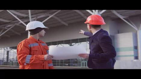 Young-manager-of-the-project-in-a-black-suit-examining-the-building-object-with-construction-worker-in-orange-uniform-and-helmet.