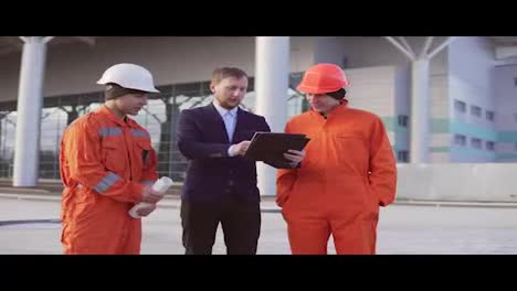 Investor-of-the-project-in-a-black-suit-examining-the-building-object-with-construction-workers-in-orange-uniform-and-helmets.-They-are-cheking-the-drawings-using-the-tablet.
