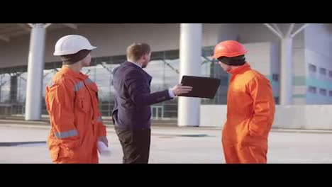 Investor-Des-Projekts-Im-Schwarzen-Anzug-Untersucht-Das-Bauobjekt-Mit-Bauarbeitern-In-Orangefarbener-Uniform-Und-Helmen.-Sie-überprüfen-Die-Zeichnungen-Mit-Dem-Tablet.