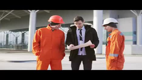 Investor-of-the-project-in-a-black-suit-examining-the-building-object-with-construction-workers-in-orange-uniform-and-helmets.-They-are-cheking-the-drawings-together.