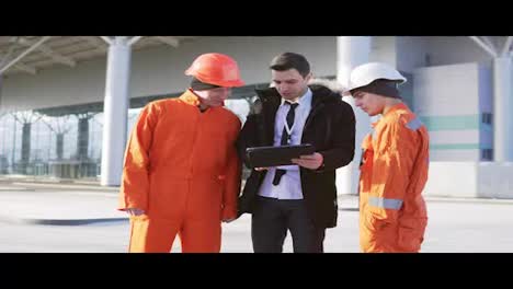 Investor-of-the-project-in-a-black-suit-examining-the-building-object-with-construction-workers-in-orange-uniform-and-helmets.-They-are-cheking-the-drawings-using-a-tablet