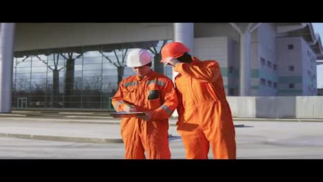 Two-construction-workers-in-orange-uniform-and-helmets-looking-over-plans-together.-Building-at-the-background