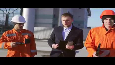 Engineer-in-a-suit-and-two-workers-in-orange-uniform-and-helmets-are-walking-through-building-facility.-Shot-in-4K