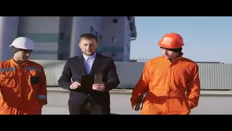 Ingenieur-Im-Anzug-Und-Zwei-Arbeiter-In-Orangefarbener-Uniform-Und-Helmen-Gehen-Durch-Die-Gebäudeanlage.-In-4k-Gedreht