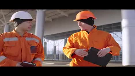 Two-construction-workers-in-orange-uniform-and-helmets-walking-through-the-construction-field-and-looking-over-plans-together.-Building-at-the-background