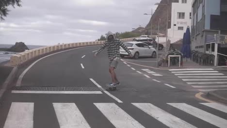 Man-longboarding-on-a-road-close-to-coastline-in-cloudy-weather.-Tall-man-in-hoodie-practicing-skateboarding-by-high-angle-road.-Slow-motion