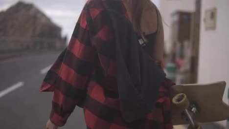 Cropped-footage-woman-in-plaid-coat-on-a-cloudy-day-at-walking,-holding-a-skateboard,-slow-motion.-Unrecognizable-girl-walks-by-asphalt-road