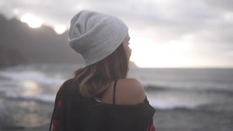 Woman-gazes-at-ocean---A-brunette-woman-shown-from-behind-looks-out-over-an-ocean-in-front-of-the-hills,-and-then-turns-her-head-to-the-side-smiling-over-her-shoulder.-Blurred-background