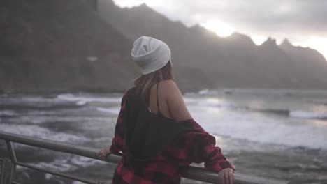 Woman-gazes-at-ocean---A-brunette-woman-shown-from-behind-looks-out-over-an-ocean-in-front-of-the-hills,-and-then-turns-and-walk-away.-Cloudy-weather.-Slow-motion