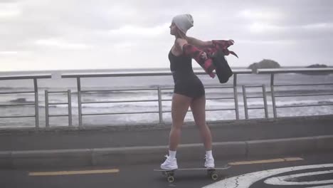 Beautiful-girl-freerly-riding-longboard-with-outstretched-hands-by-the-coastline-road-in-cloudy-weather.-Portrait-hipster-girl-smiling-with-a-longboard-at-sunset.-Slow-motion.-Side-view