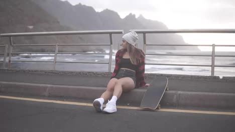 Girl-sitting-with-longboard-at-the-border-at-coastline-asphalt-road-on-background-hills-and-foggy-ocean.-Relaxing-break.-Cloudy-weather