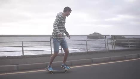 Sport,-lifestyle,-extreme-and-people-concept---Tall-guy-riding-longboard-by-the-coastline-road-in-cloudy-weather.-Full-length-of-an-active-man-exercising-on-road.-Slow-motion