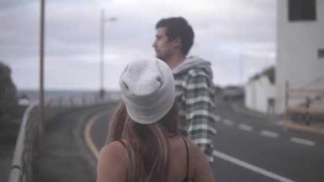Thoughtful-man-gazes-at-ocean---Young-couple-shown-from-the-side-standing-on-coastline-road.-Cute,-stylish-girl-comes-to-her-boyfriend-to-kiss-him.-Cloudy-weather