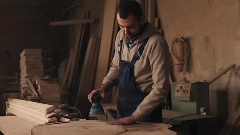 A-carpentry-shop-with-wood-material,-tools-on-the-background.-Joiner-working-on-the-processing-of-wood-in-blue-overalls.-Carefully-grinds-the-material-with-a-hand-held-machine