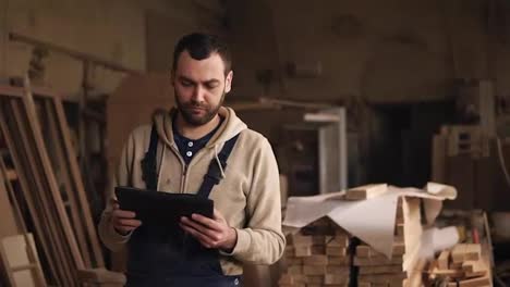 A-young-man-with-a-beard-walks-around-the-carpentry-shop-with-a-tablet-in-his-hands.-Looks-back,-evaluates