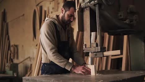 Carpenter-shop.-A-nice-worker-cuts-a-ribbed-shape-from-a-wooden-block-using-an-electric-saw-machine.-Evaluates-the-work,-holding-the-product-in-hands