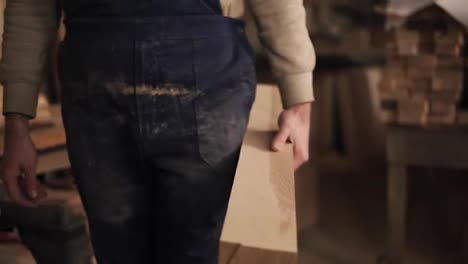 Slow-motion-footage-of-a-young-carpenter-with-rough-hands.-A-man-walks-in-a-dirty-with-wood-dust-blue-overalls-with-a-bar-of-wood-in-his-hands