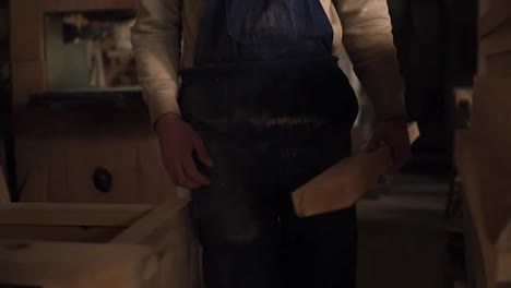 The-carpenter-in-the-workshop-goes-to-the-grinding-machine-with-a-bar-of-wood.-A-man-in-blue-overalls.-Its-dusty-from-working-with-a-wood-.-Without-a-face