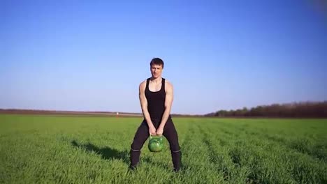 A-strong-man-demonstrates-an-exercise-with-weight.-Lifting-the-bob-with-both-hands.-Clear-blue-sky,-green-field.-Front-footage