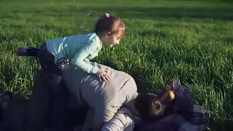 Wide-meadow-with-green-grass.-Young-father-and-two-daughters-are-having-fun-together.-Fool-around.-The-youngest-has-jumped-on-the-fathers-back.-Playing-on-the-ground,-on-a-picnic-blanket