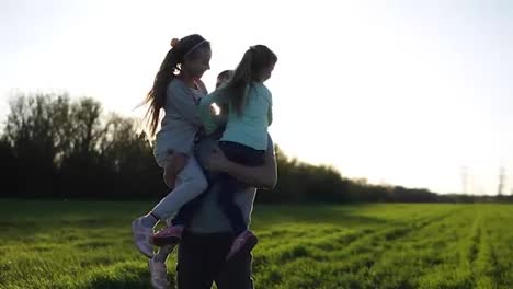 Proud-father-of-two-girls-in-nature.-He-carries-them-in-his-arms,-the-sun-is-shining,-the-youngest-girl-sends-air-kisses.-Beautiful-field-with-green-grass.-Slow-motion