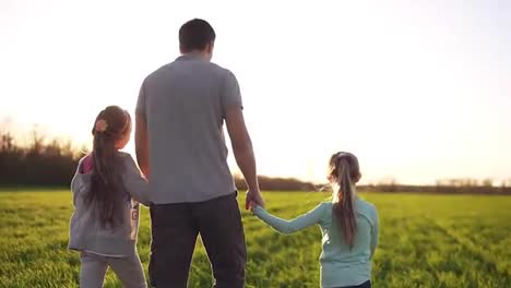 Young,-muscular-man,-father-is-holding-his-small-daughters-with-hands.-Walking-by-the-wide-meadow.-Casual-clothes.-Sun-rays.-Footage-from-the-backside