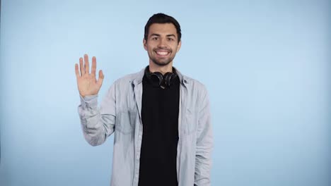 Cropped-shot-of-happy-excited-handsome-caucasian-man-in-blue-shirt-standing,-looking,-toothy-smile,-surprised-and-waving-his-hand-and-greeting.-Indoor-studio-shot-isolated-on-blue-wall-background