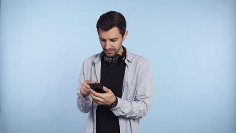 Handsome,-curious-man-in-blue-t-shirt-and-headphones-on-neck-presenting-smart-phone-and-showing-green-screen-smartphone-to-the-camera,-isolated-on-blue-background