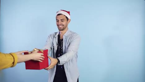 Positive,-young-guy-in-Christmas-hat-and-garland-on-neck-recieves-surprise-from-from-the-side-female-hands---being-in-good-festive-mood,-isolated-over-blue-background.-People,-surprisment-and-presents-concept