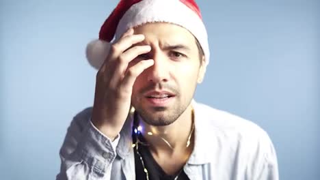 Closeup-footage-of-young-attractive-man-in-santa-claus-red-hat-after-celebration-or-long-night-looking-at-camera-mirror-and-touching-his-face,-recognizing-himself,-preen.-Background-isolated-on-blue