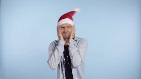 Young-crazy-guy-in-traditional-santa-hat-shaking,-waving-head-head,-getting-crazy---holding-hands-on-face,-grimace-while-standing-isolated-over-blue-background