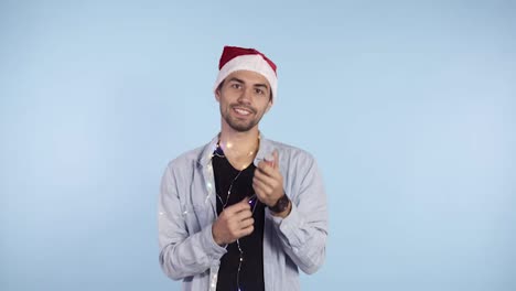 Young,-smiling-man-in-casual-clothes-and-santa-hat-on-a-head-exploding-confetti-cracker-on-a-blue-background.-Happy-celebrating-of-a-New-Year-or-Chrismas