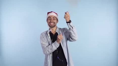 Young,-smiling-man-in-casual-clothes-and-santa-hat-on-a-head-exploding-confetti-cracker-on-a-blue-background-and-starting-to-jump-in-excitement.-Happy-celebrating-of-a-New-Year-or-Chrismas