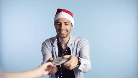 Shocked-man-received-paper-money,-dollar-banknotes.-Counting-money-while-standing-against-the-blue-wall-in-santa-hat-and-garland-on-neck.-Get-money-cash,-receive-winner-payment,-gift-concept.-Man's-hand-hold-and-counting-cash-money-and-showing-as-fan-in-excitement