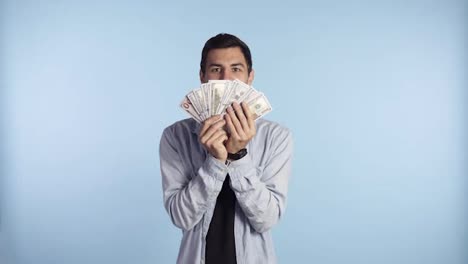 Astonished-caucasian-man-in-casual-clothes-holding-bunches-of-dollars,-lucky-lottery-winner,-super-excited-about-it.-Holding-paper-money-before-face,-can't-believe-in-this-luck.-Blue-wall-background