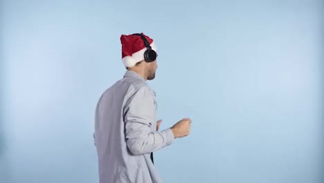 Positive-handsome-man-in-a-Christmas-red-hat-having-headphones,-listening-to-his-favorite-music-and-dances-in-a-carefree-rhythm--blue-wall-background-studio.-Man-in-casual-clothes-and-hat-freerly-dances-in-front-the-camera