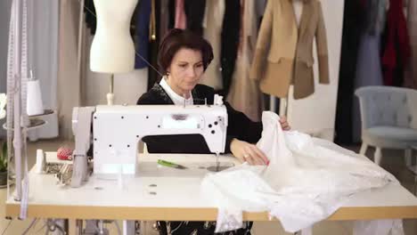 Portrait-of-female-tailor-working-on-sewing-machine.-Caucasian-woman-using-a-sewing-machine-at-studio-or-in-workshop.-Skillful-expert-tailor-works-on-sew-machine-with-a-white-pattern,-sparkling-fabric