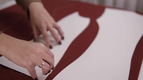 Female-tailor-is-working-at-table-with-red-cloth-in-atelier.-Skilled-seamstress-is-drawing-sketch-lines-with-a-soap-or-a-chalk-on-red-fabric-using-patterns.-Close-up-of-hands-with-perfect-manicure.-Slow-motion
