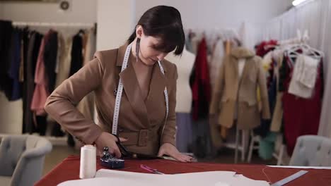 Female-fashion-designer-in-stylish-brown-coat-and-earrings-working-with-red-fabric-at-the-studio-full-of-tailoring-tools---pattern,-scissors,-measuring-tape.-Workplace-of-seamstress.-Tailor-cuts-future-dress-detail-then-cheerfully-smiling-to-the-camera