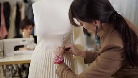 Creative-fashion-designers-are-working-together-in-the-tailor-studio.-Long-haired-woman-pinning-on-the-white-skirt-on-mannequin-using-pins-on-her-hand-pillow-on-wrist.-Woman-working-on-sewing-machine-on-a-blurred-background