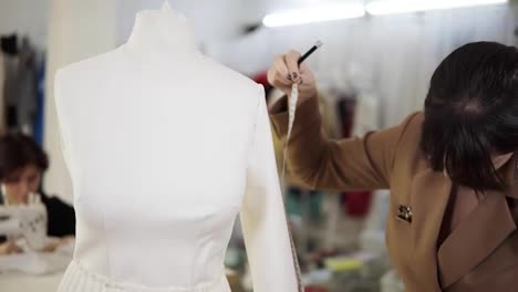 Modern-sewing-studio,-concentrated-brunette-female-fashion-designer-measuring-sleeve-from-a-white-suit-on-mannequin-and-making-notes-holding-her-notebook.-Slow-motion